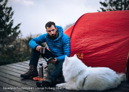 Ein Wanderer sitzt auf einem Trekkingplatz in Hallenberg mit seinem Hund vor einem Zelt
